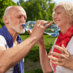 Older man and woman smiling and sharing a water bottle in a sunny park.